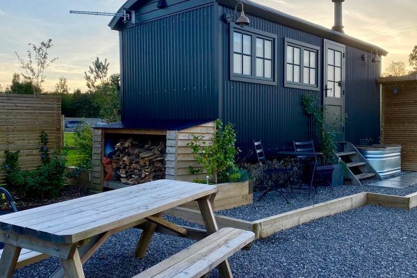 Dark blue modern cabin with picnic table, gravel patio, stone fire pit, and water feature at dusk