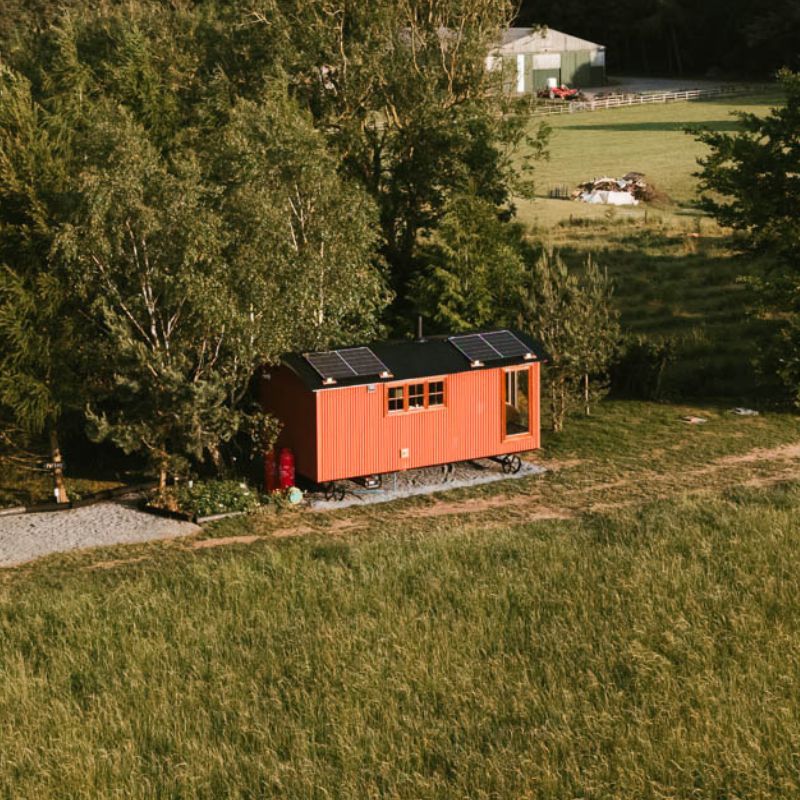 Orange tiny house with solar panels beside trees in a grassy rural field