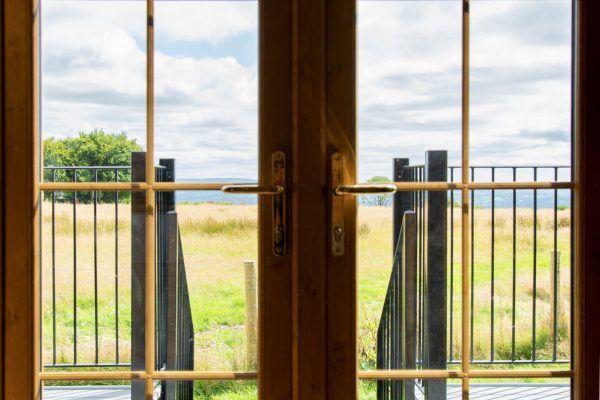 Wooden French doors open to a patio and grassy field with black railings under a cloudy sky