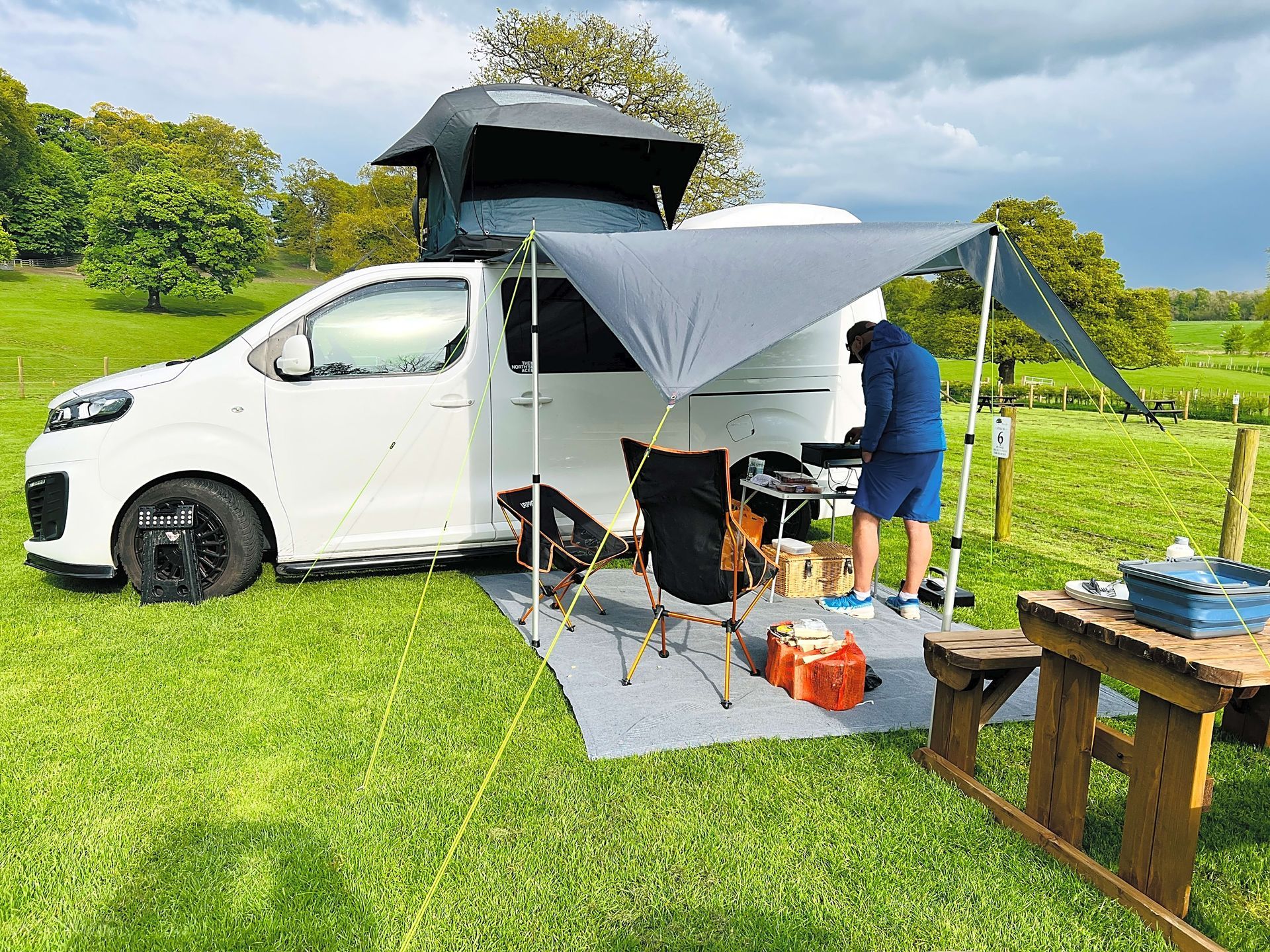 White camper van with rooftop tent and awning set up in a grassy campsite, with camping gear nearby.