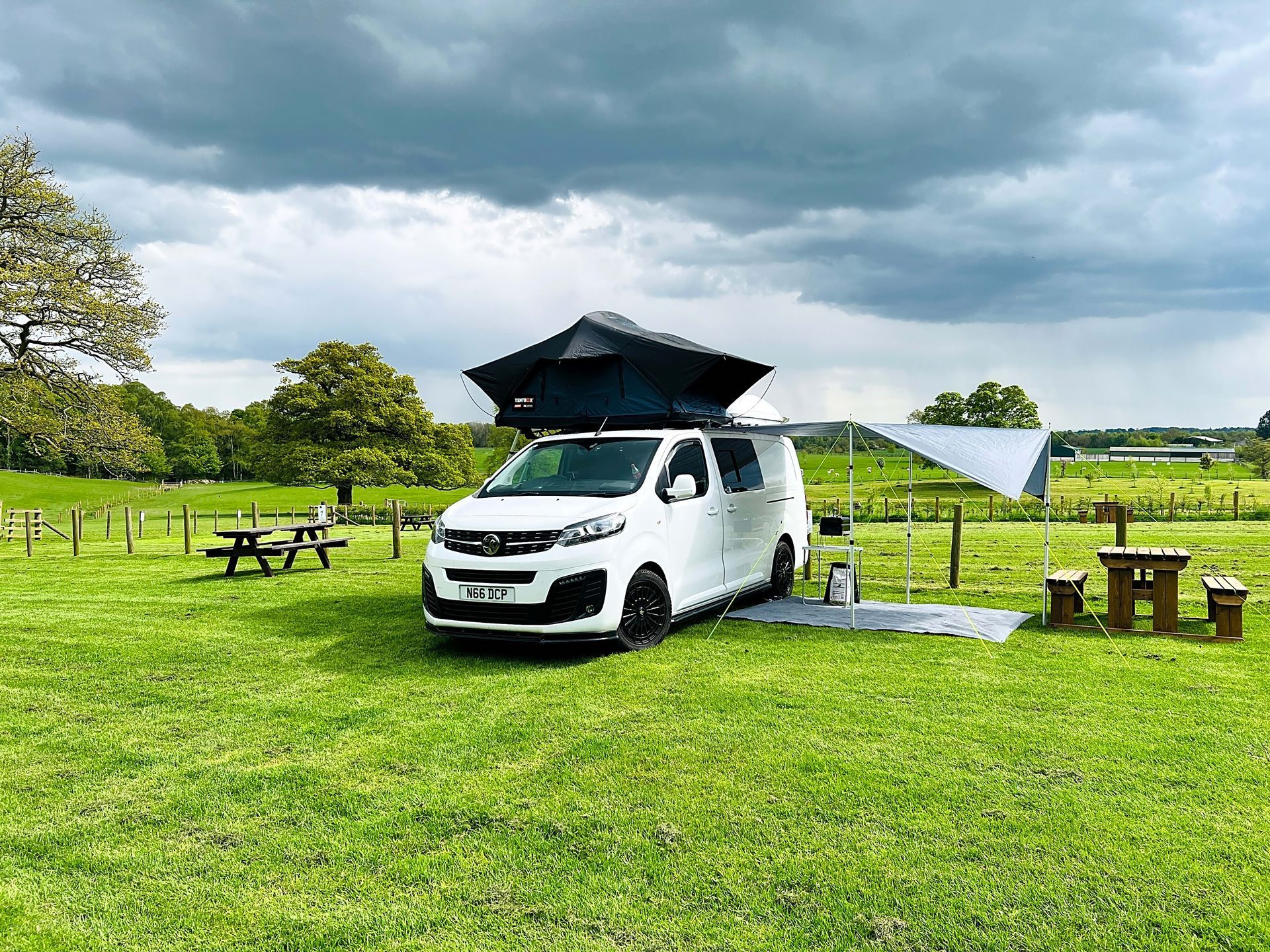 White van with open rear hatch at a grassy campsite under cloudy skies, with picnic tables and tents nearby