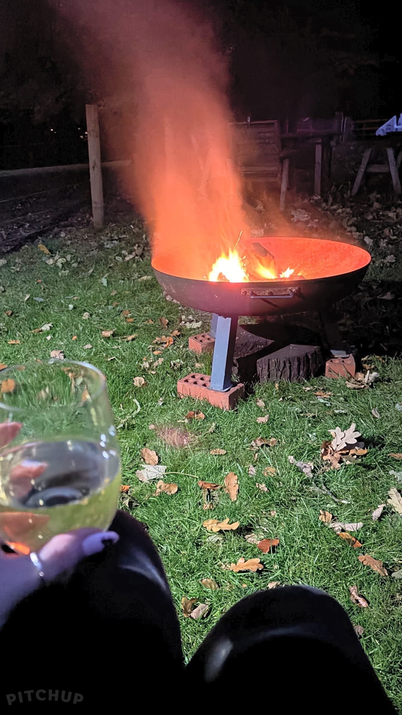 Person holding a drink beside a glowing fire pit in a leaf-covered backyard at night