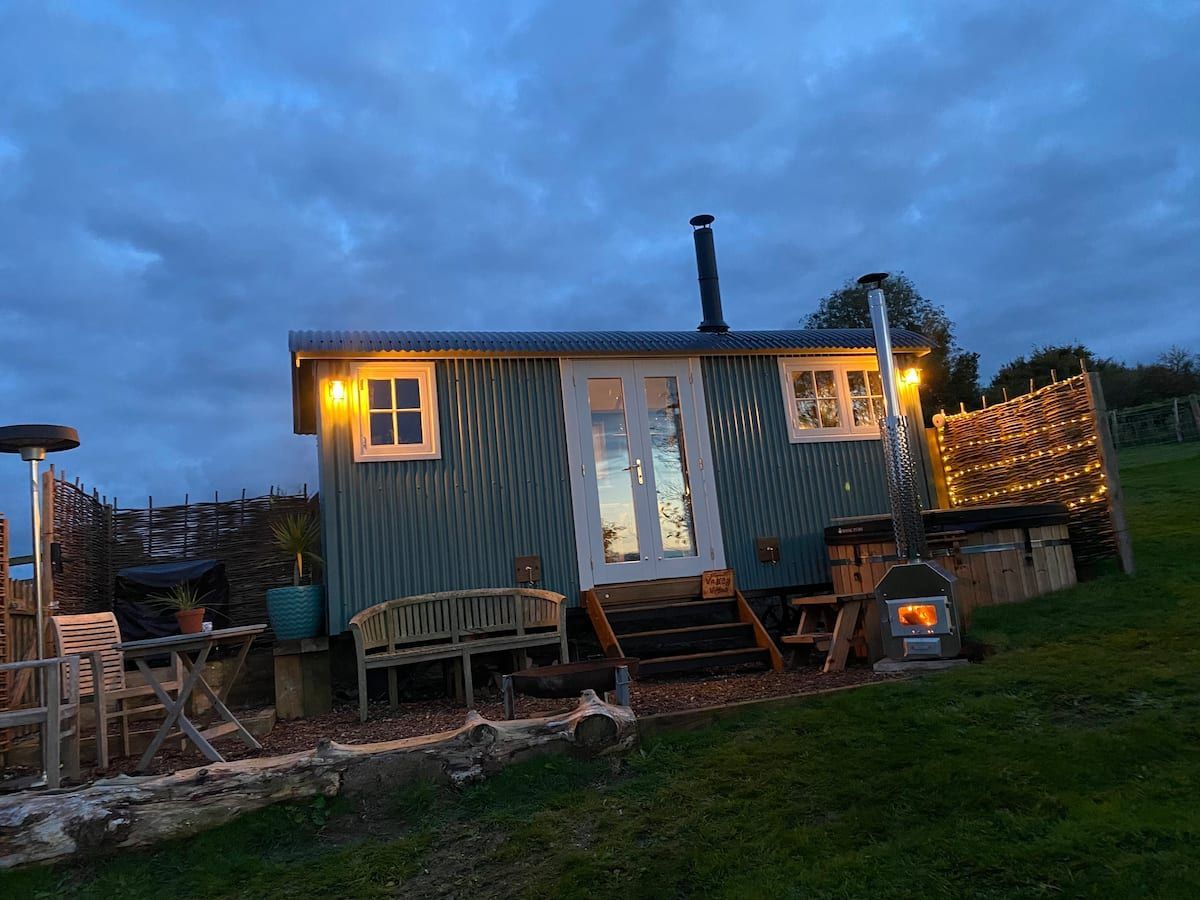 Small teal cabin with lit windows and string lights at dusk, beside a grassy yard and stone patio