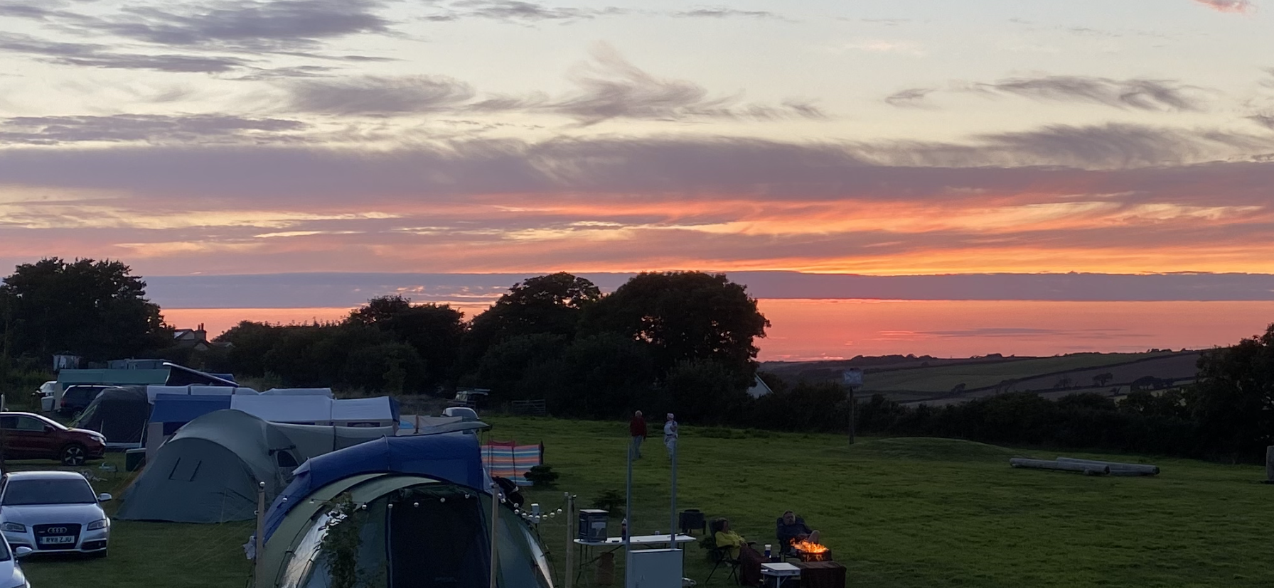 Sunset over a grassy campsite with tents, trees, and parked cars under a colorful sky