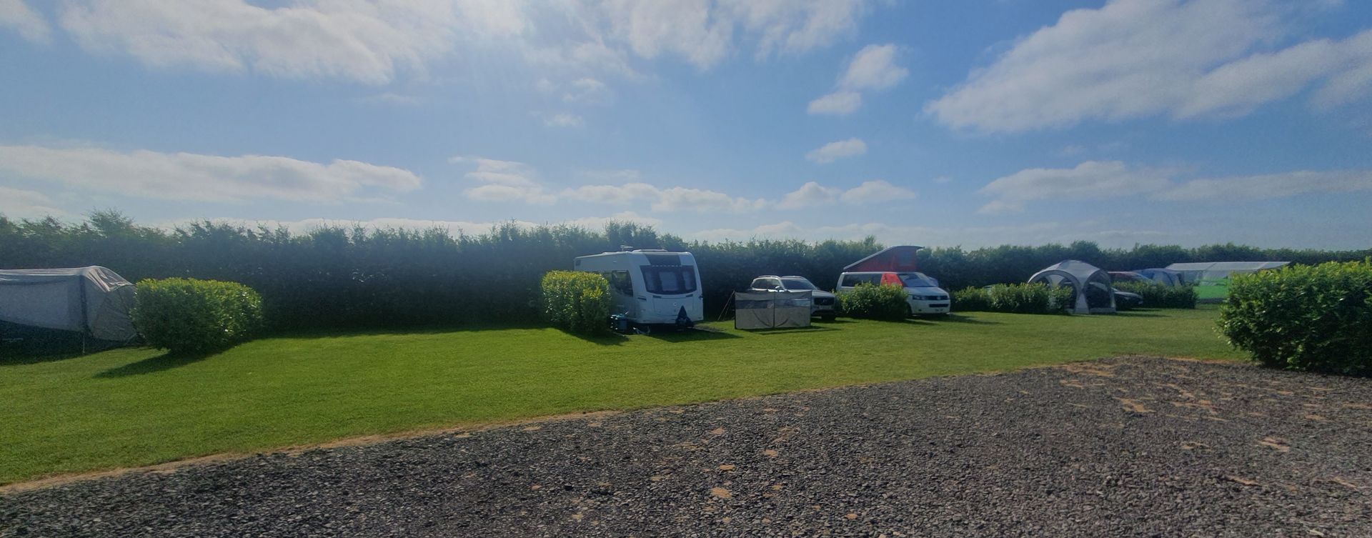 Wide grassy field with parked campers under a cloudy sky, gravel in the foreground