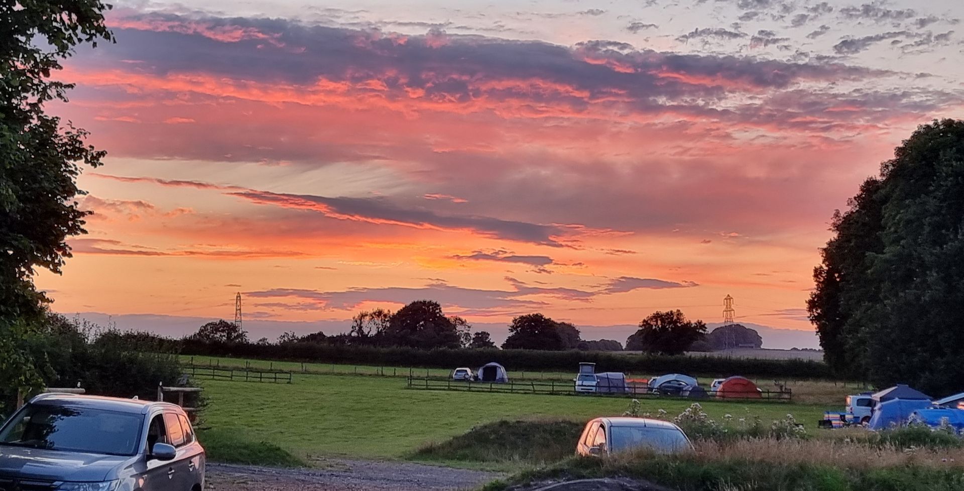 Sunset over a grassy campsite with parked cars and tents under a pink-orange sky