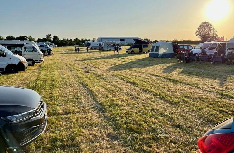 Camping area with tents, parked cars, and an airplane in the distance at sunset