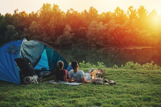People relaxing by a blue tent beside a lake at sunset, with food on a picnic blanket