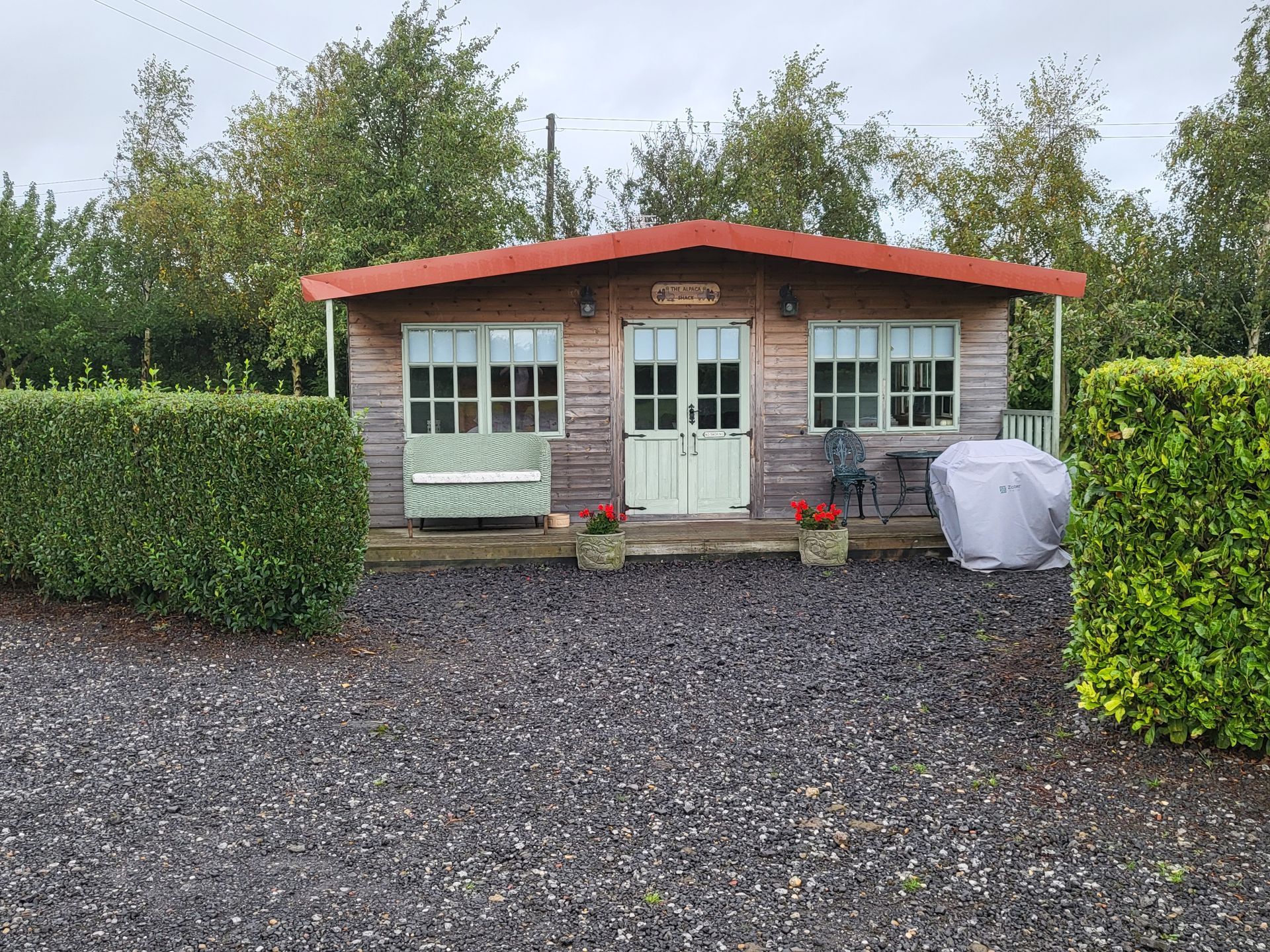 Small garden shed with a red roof, white doors, and windows, set behind a gravel yard and hedges.