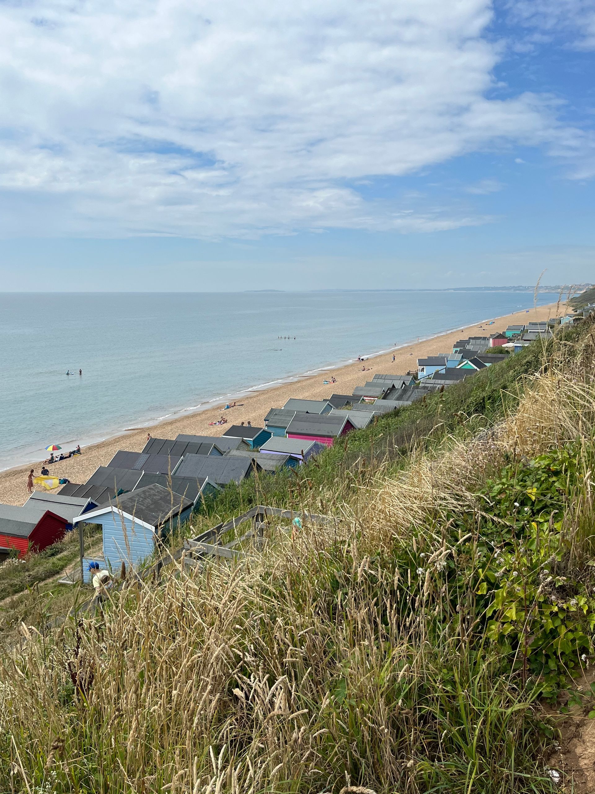 Sunny beach coastline with calm sea, sandy shore, and colorful rooftops below a grassy hillside.