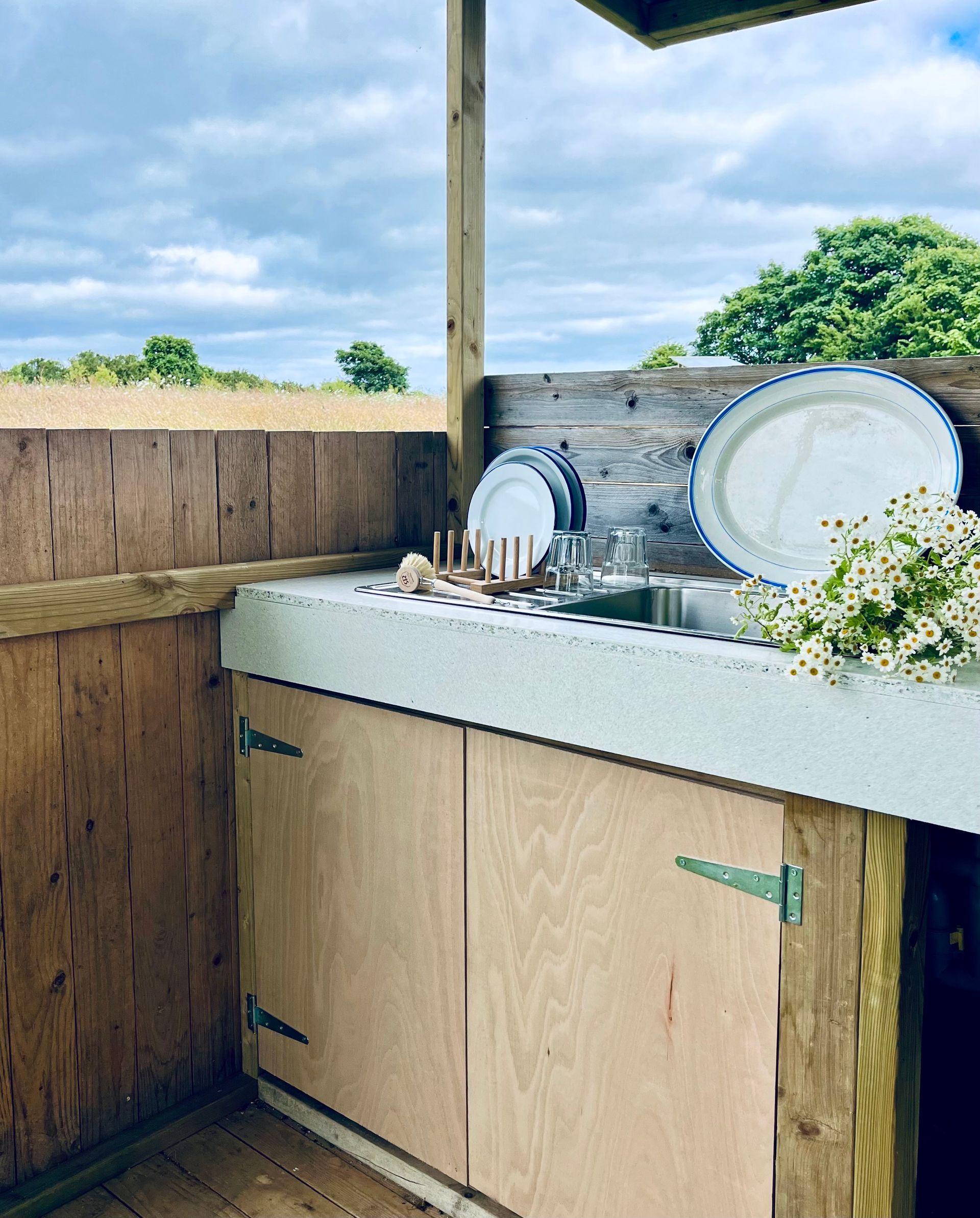 Outdoor kitchen with wooden cabinets, stone countertop, stacked dishes, and flowers under a patio roof