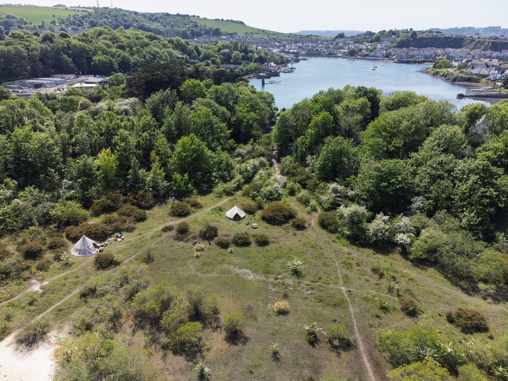 Aerial view of a green hillside with tents overlooking a river or lake and dense forest.