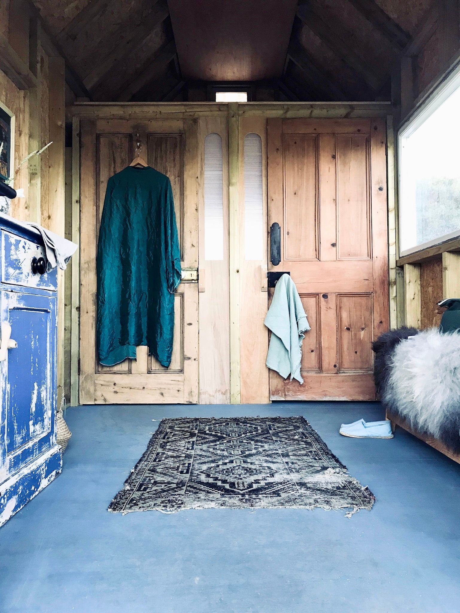 Wooden porch with blue floor, hanging coats, patterned rug, and a furry bench by the door