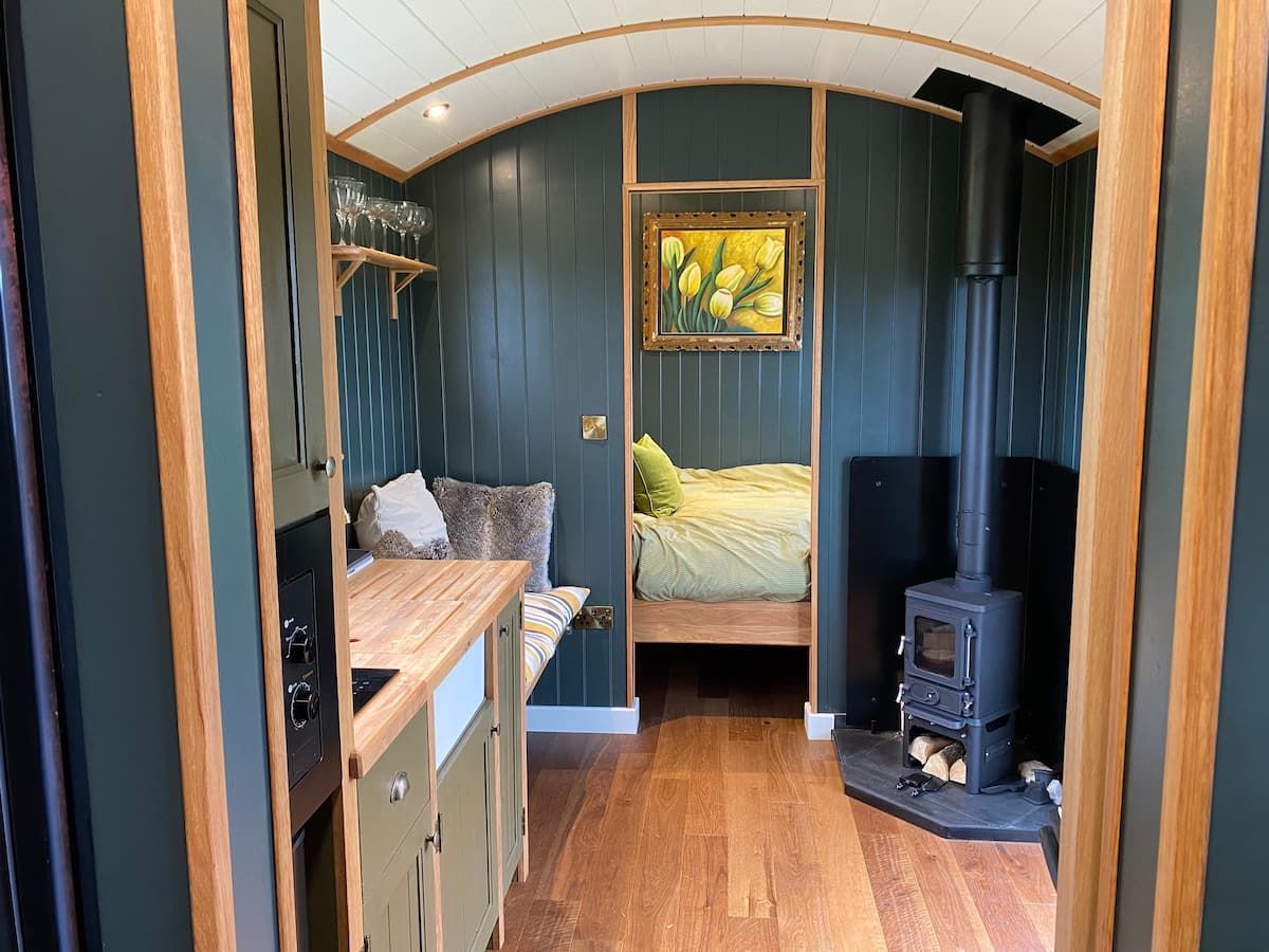 Narrow cozy cabin interior with blue paneling, wooden floor, bed, desk, and small stove.