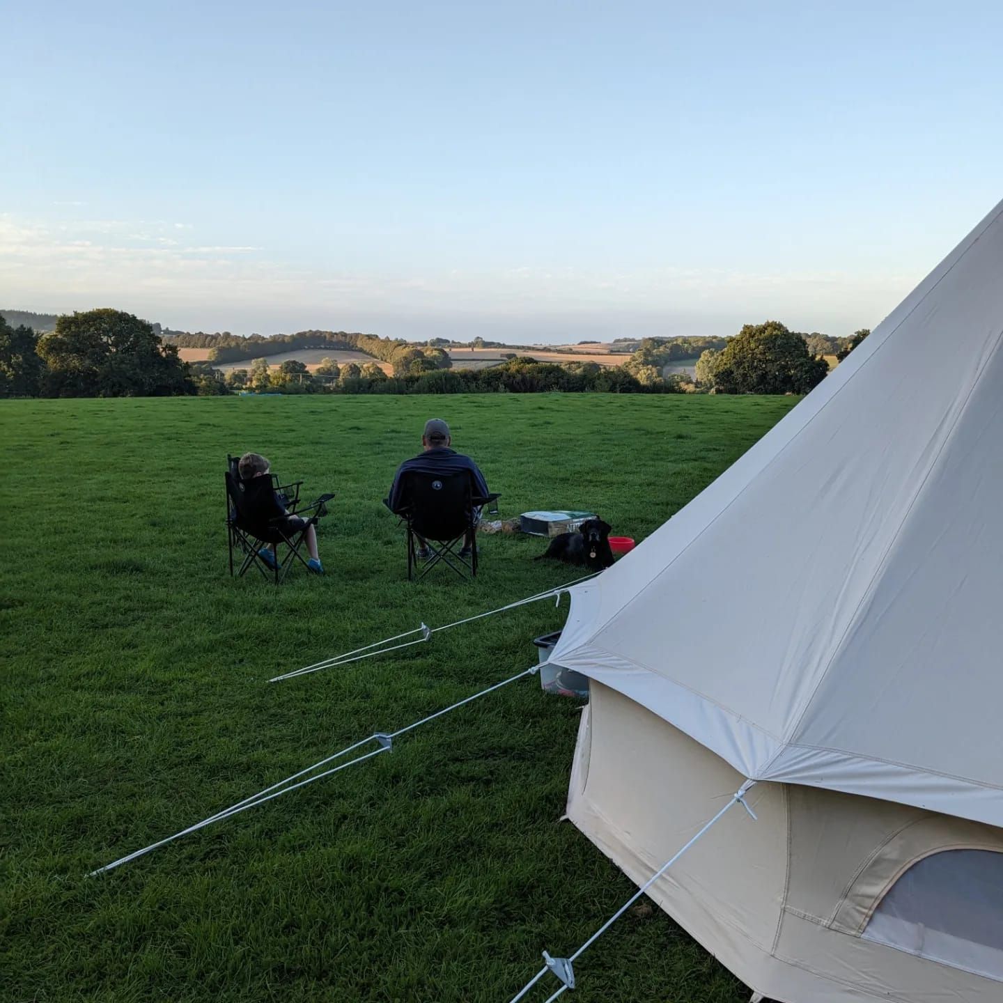 Two people sit in folding chairs on a grassy field beside a white tent at sunset.