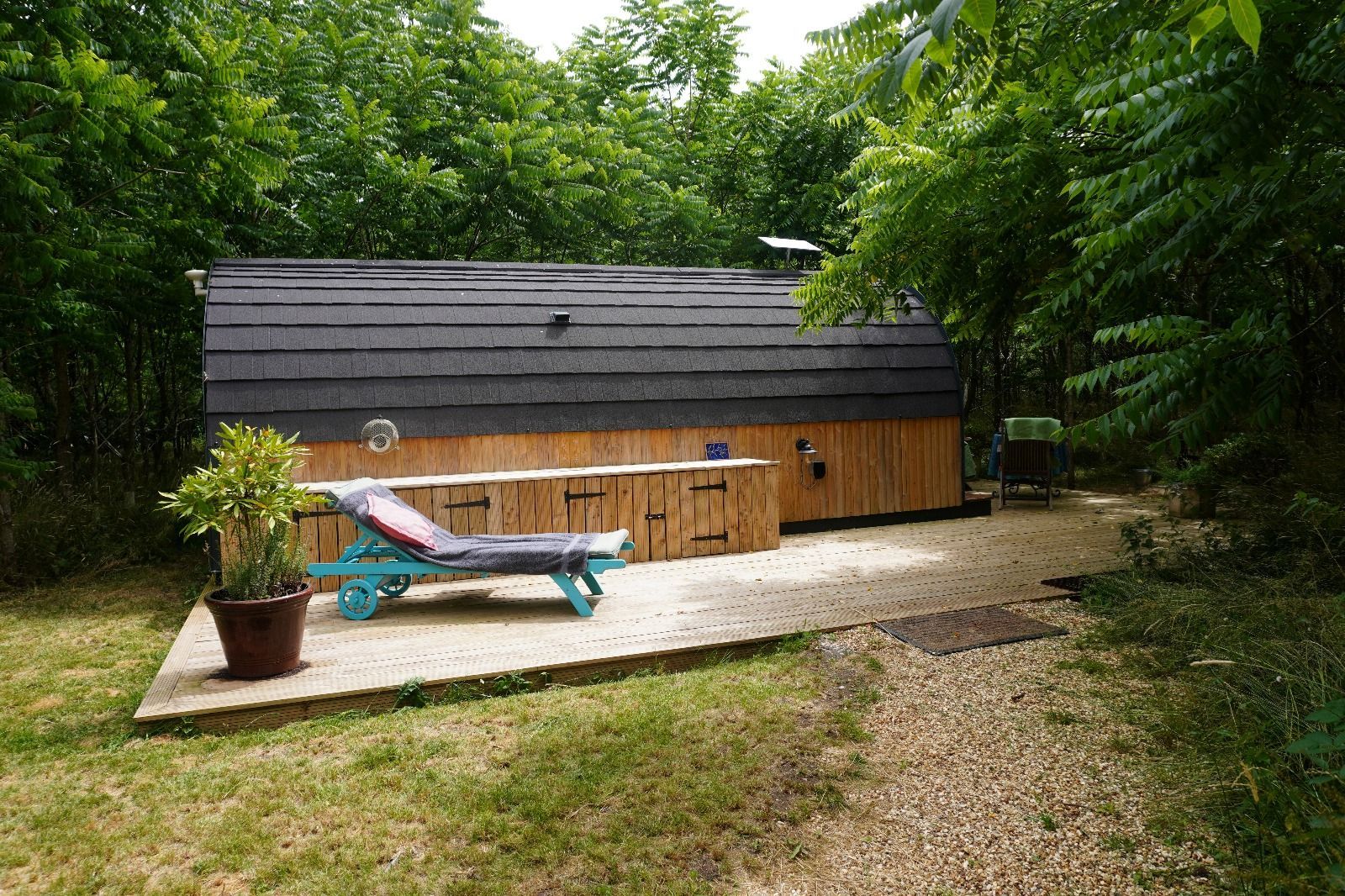 Backyard hot tub beside a wooden cabin, with a lounge chair and potted plant on a gravel patio.