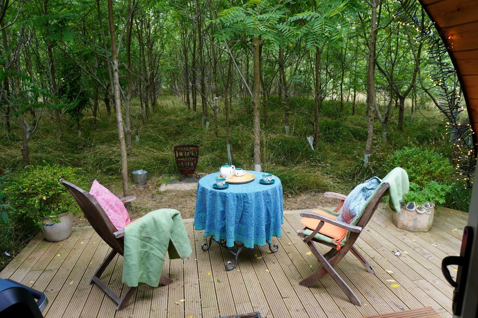 Wooden deck in a wooded garden with two chairs, a blue table, and blankets draped over the chairs.