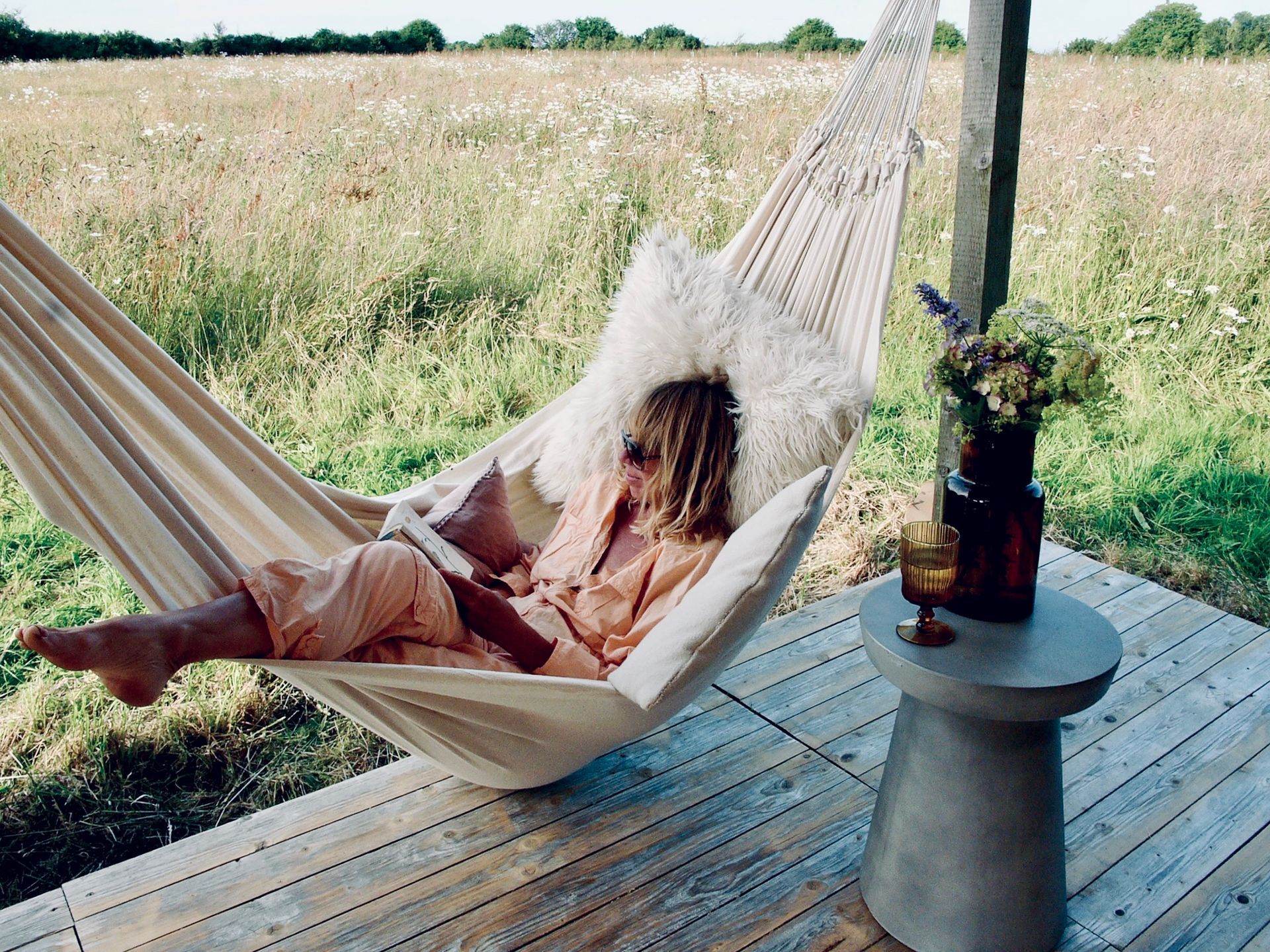 Person relaxing in a hammock on a wooden deck beside a field, with a small table and flowers nearby.