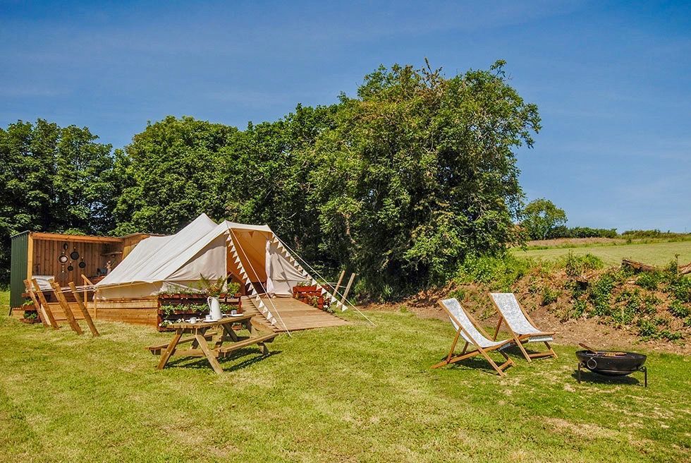 Glamping tent on grassy field with picnic table and lounge chairs under a blue sky