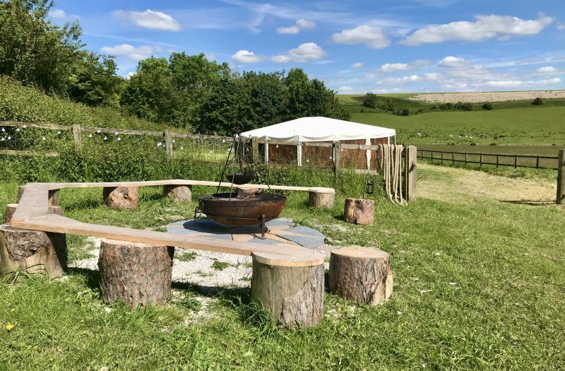 Outdoor fire pit with log benches on a grassy field beside a white canopy and wooden fence
