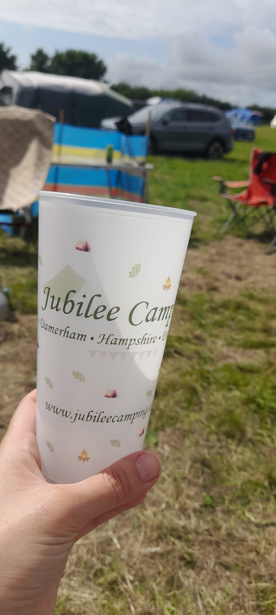 Hand holding a white “Jubilee Cup” cup outdoors near parked cars and a blue canopy.
