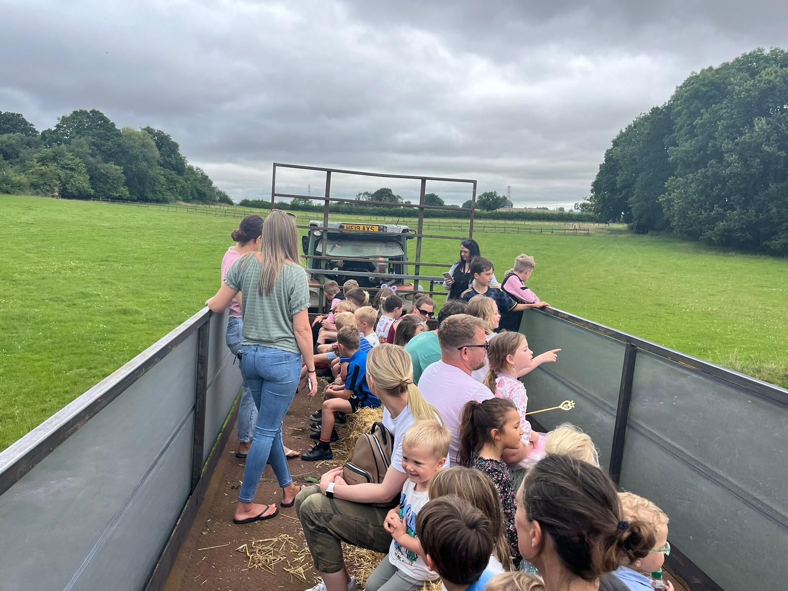 Children riding in an open trailer on a farm, with an adult and green fields under a cloudy sky.