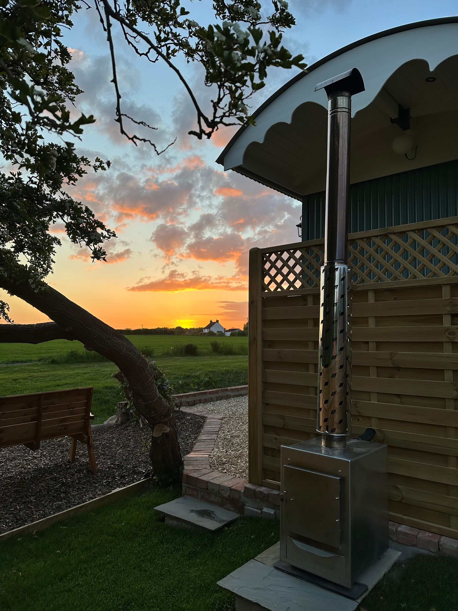 Patio heater beside a wooden deck at sunset, with a tree framing the glowing sky over fields.