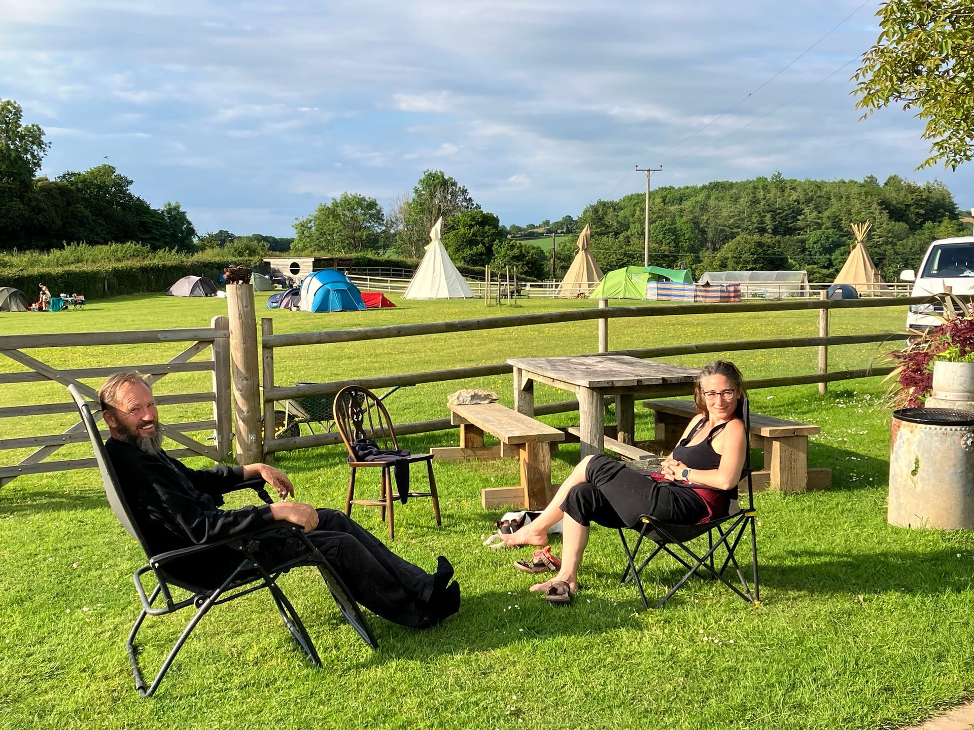 Two people relaxing in lawn chairs at a sunny campsite with tents and picnic tables nearby