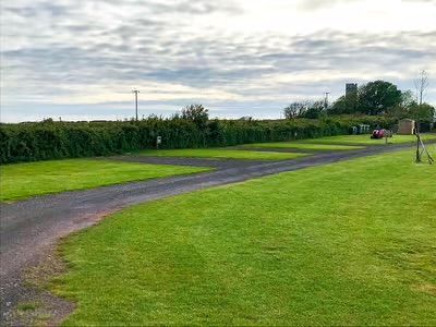 Gravel path through a green field toward hedges under a cloudy sky, with a distant house and trees.