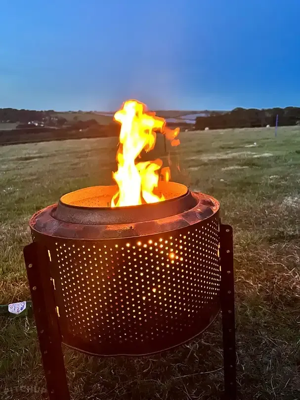 Outdoor metal fire pit with bright flames burning in a grassy field at dusk
