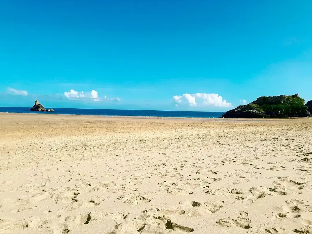 Wide sandy beach under a bright blue sky, with calm sea and a small green island in the distance.