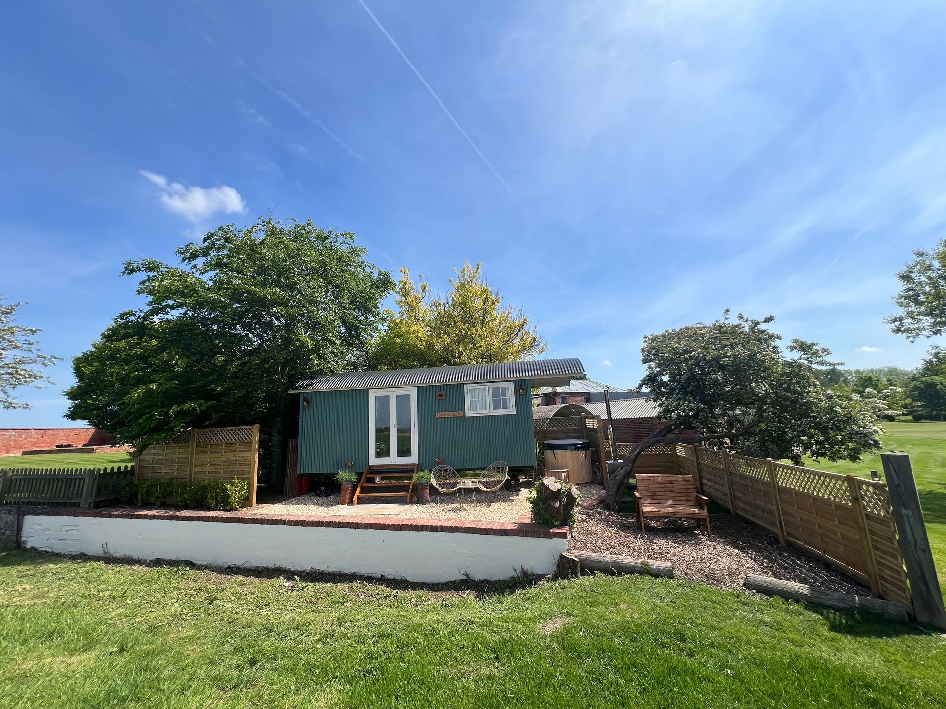 Small green cottage with a white wall, trees, and a fenced yard under a bright blue sky.