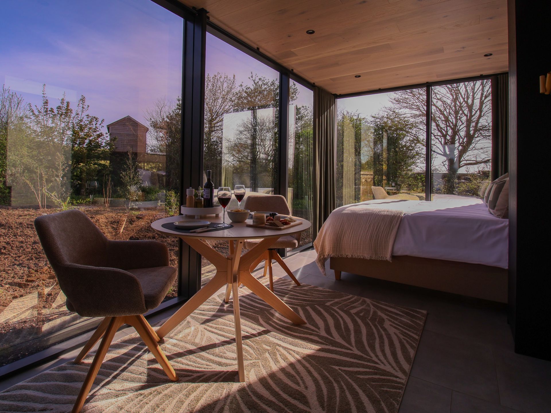 Modern bedroom with floor-to-ceiling windows, a small table and chair, and a forest view at sunset