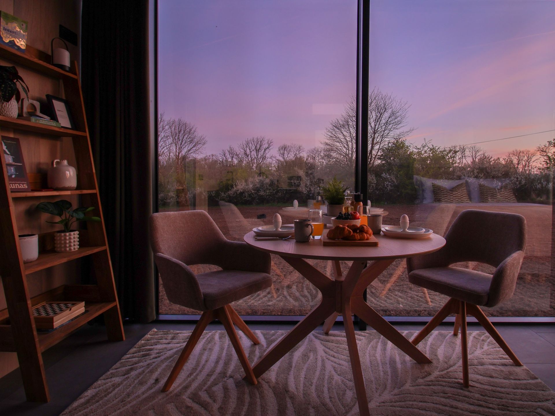 Cozy dining nook by a large window at sunset, with a round table, chairs, and soft pink sky views