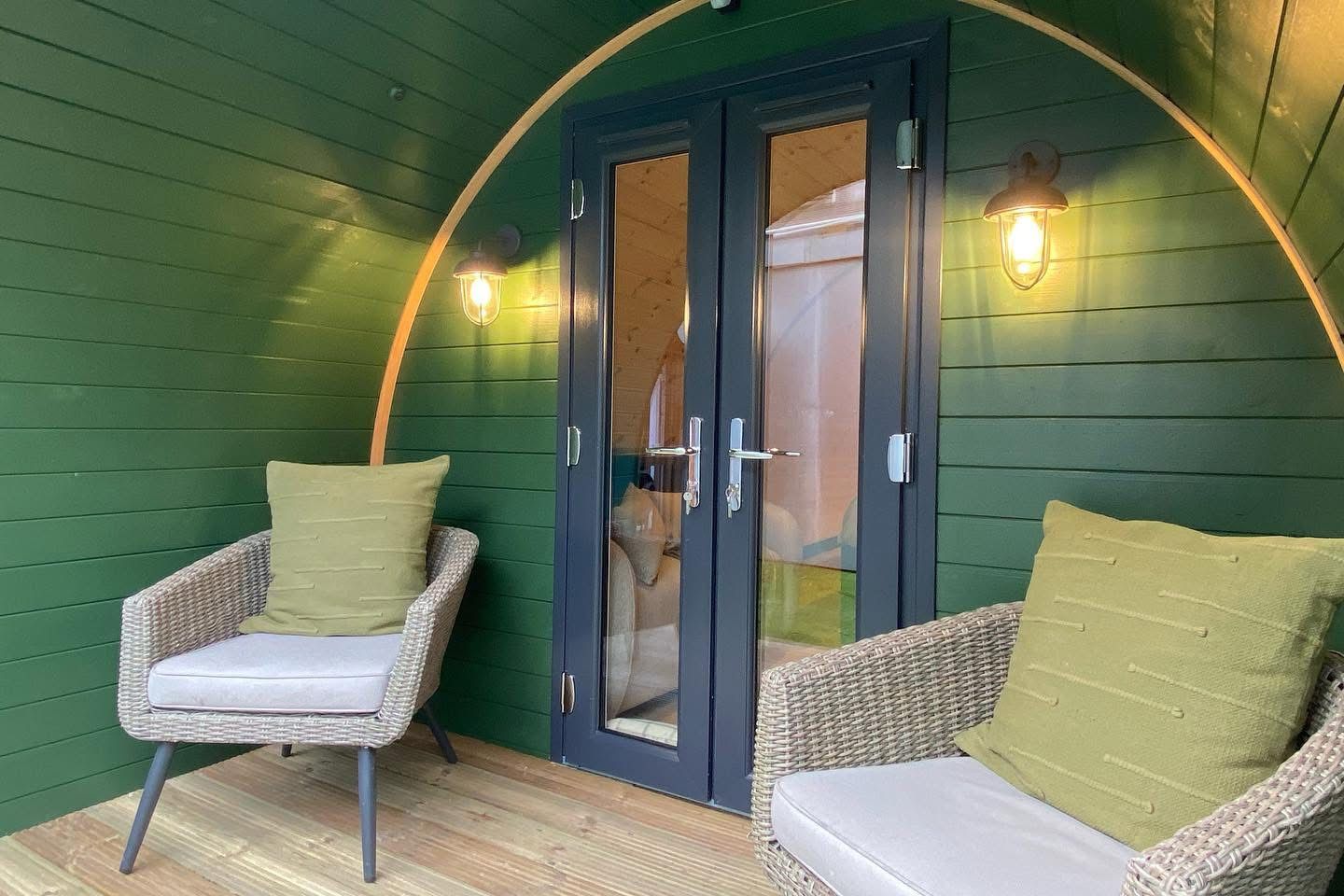 Green porch with double glass doors, wall lights, and two wicker chairs with cushions.