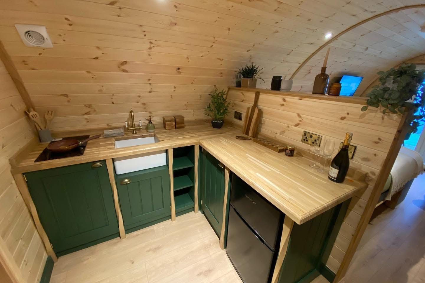 Wood-paneled tiny kitchen with green cabinets, wood counters, sink, and mini fridge under an arched ceiling