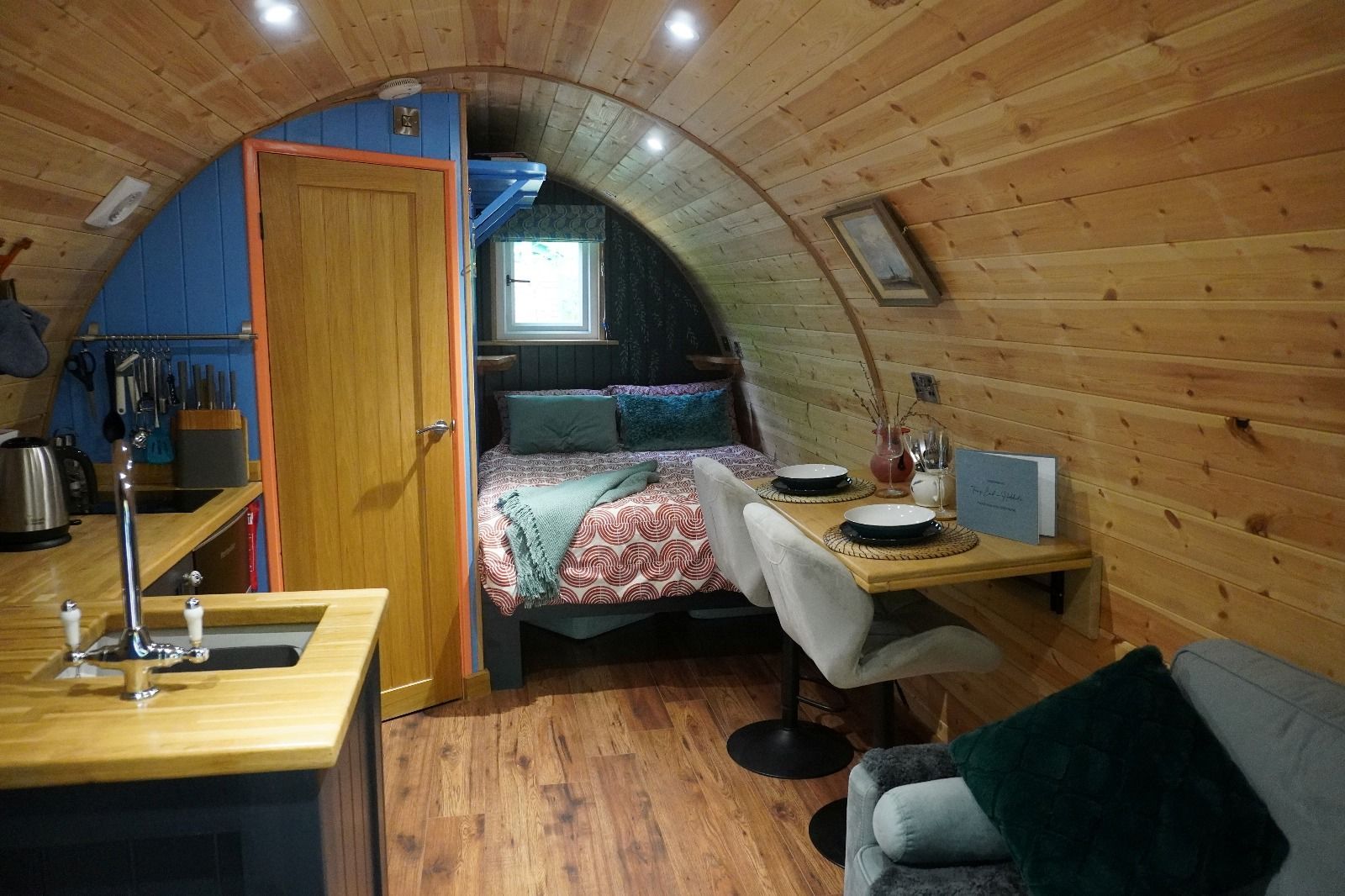 Cozy wood-paneled tiny cabin with a bed, desk, and kitchenette under a curved ceiling.