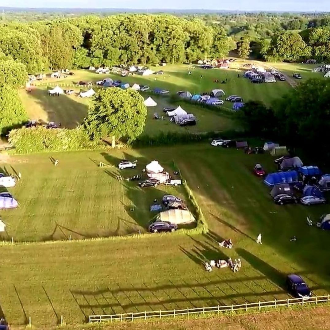 Aerial view of a grassy campsite with tents and cars beside a wooded area in evening light
