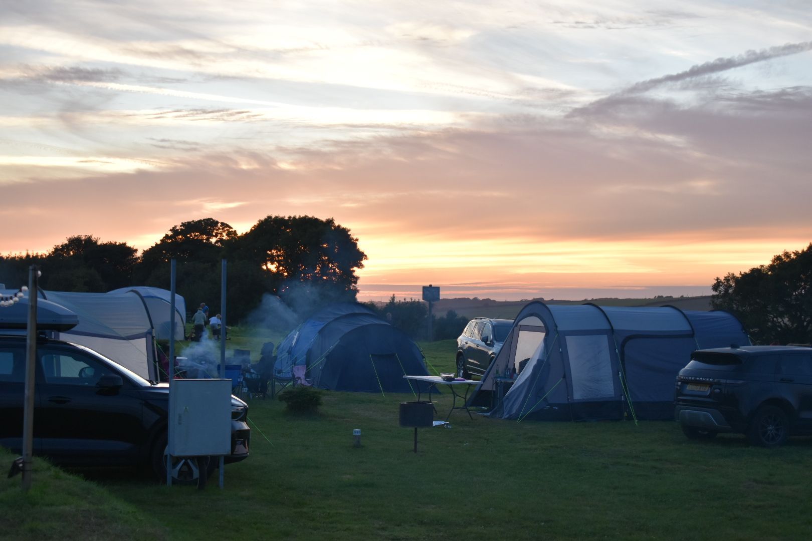 Campsite at sunset with tents, parked cars, and a glowing sky over a grassy field