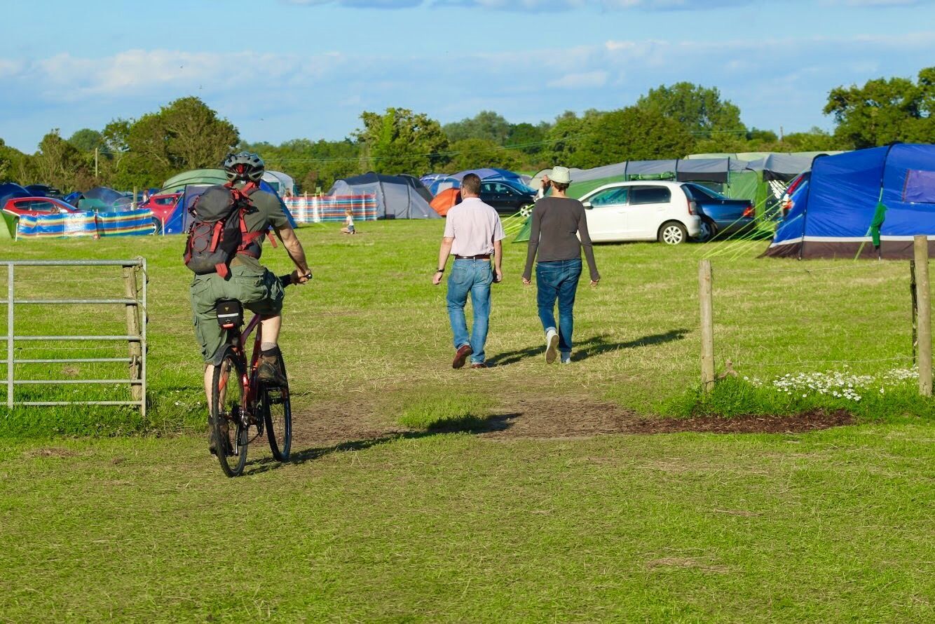 Cyclist riding past tents and parked cars in a grassy field on a sunny day