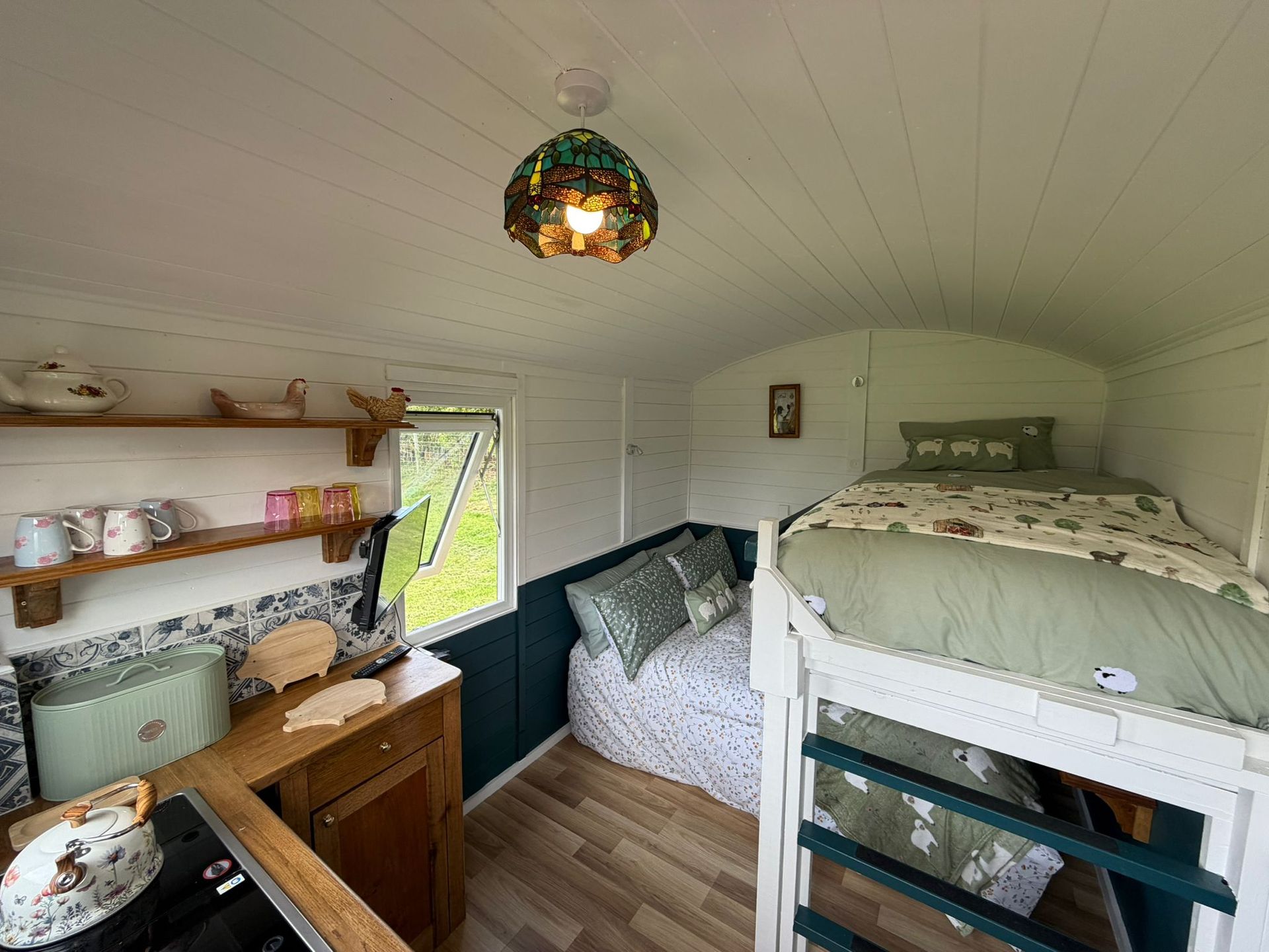 Small white bedroom with bunk beds, floral bedding, shelves, and a wood desk under a stained-glass ceiling light