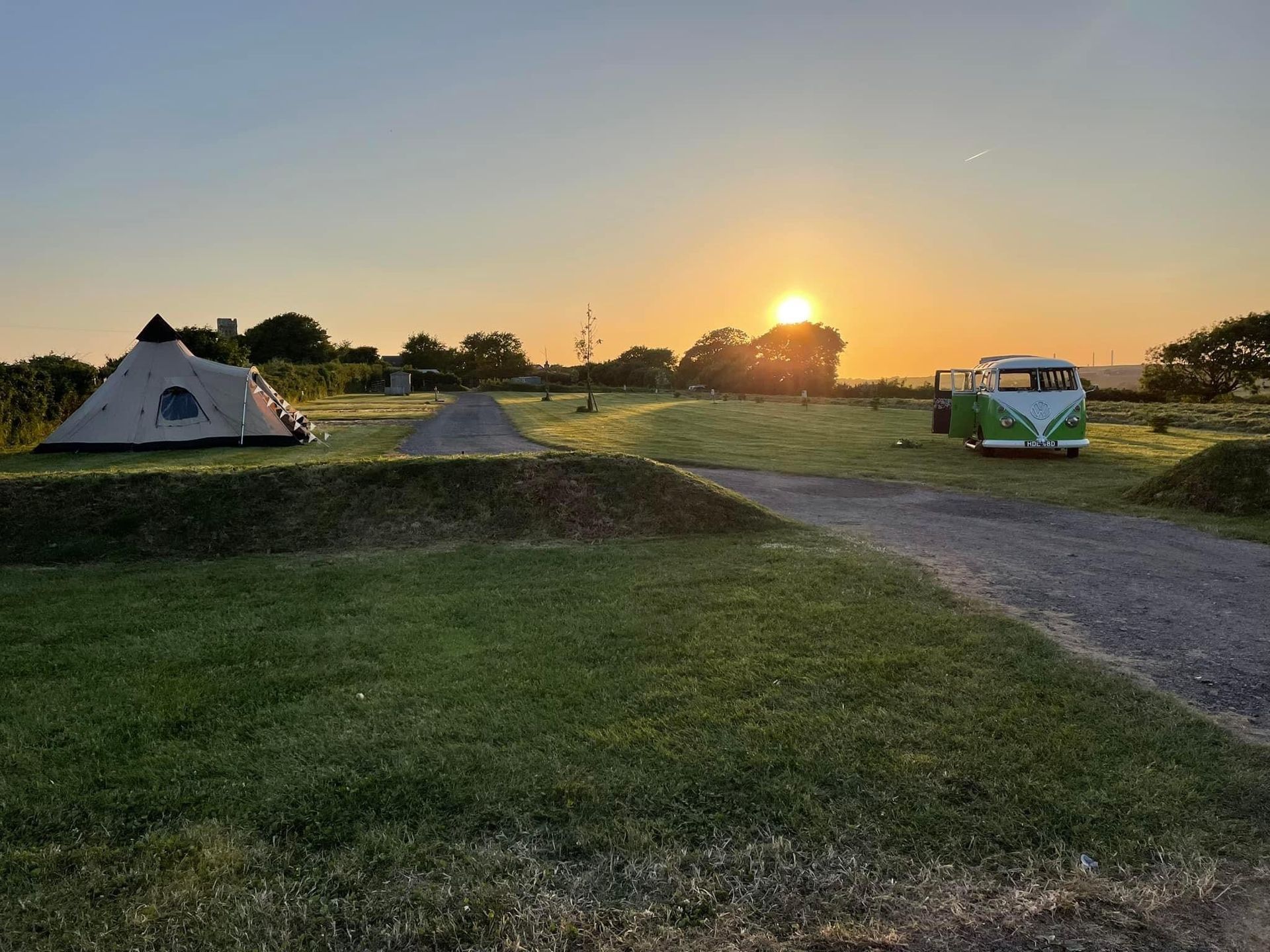 Sunset over a grassy campsite with tents, a gravel path, and a parked van