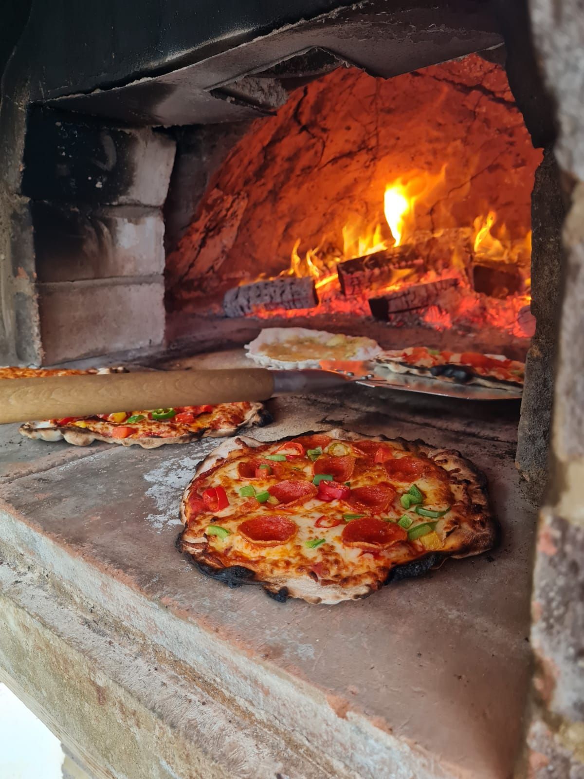 Two pizzas baking in a wood-fired oven, with flames glowing inside.