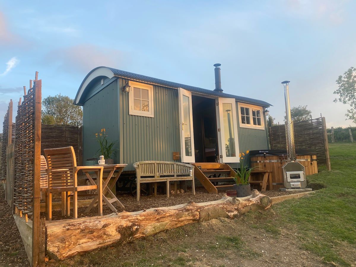 Small green cabin with deck chairs and table on a grassy rural lot at sunset