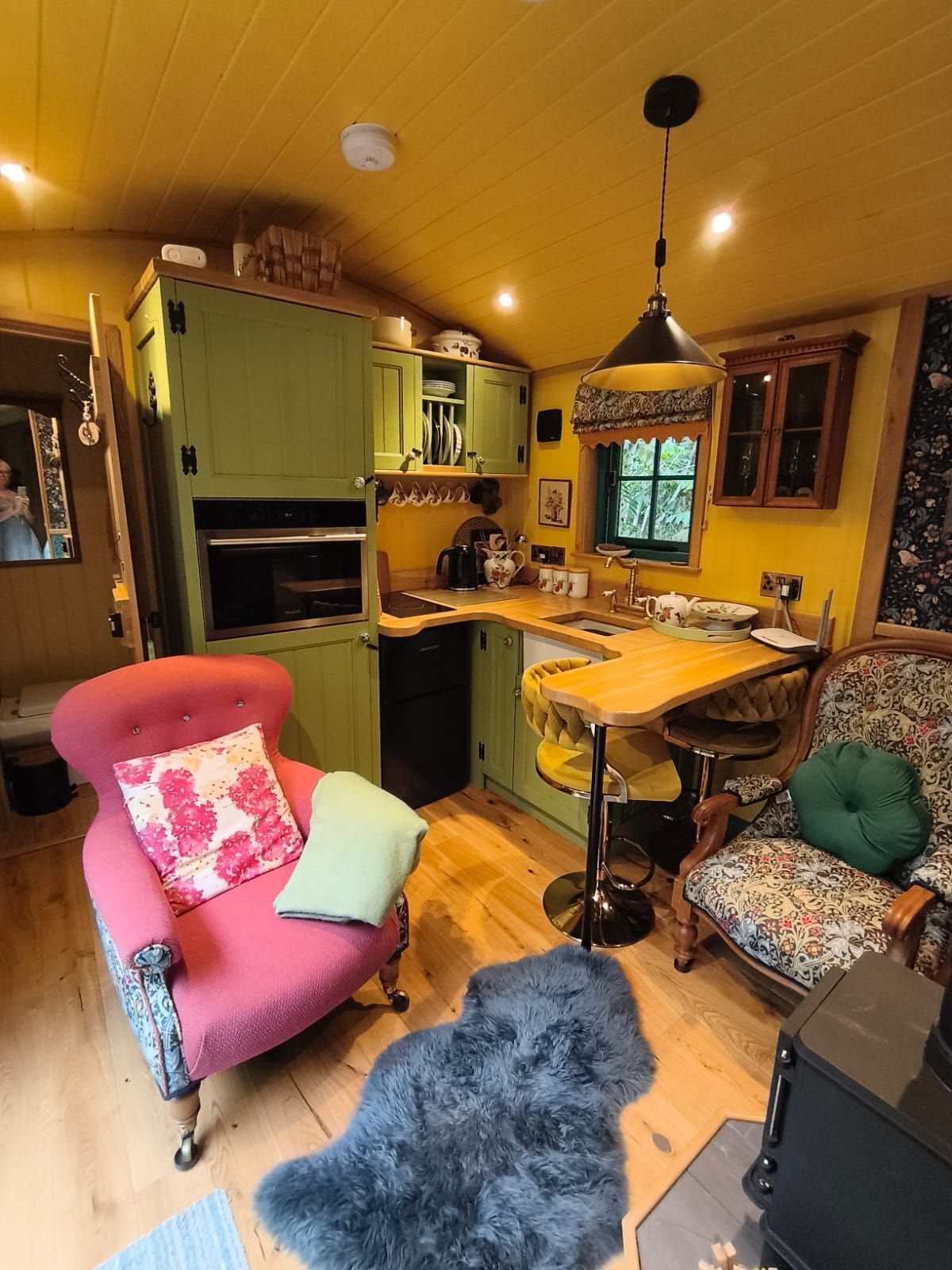 Cozy yellow kitchen-living room with colorful armchairs, wooden table, green cabinets, and hanging pendant light