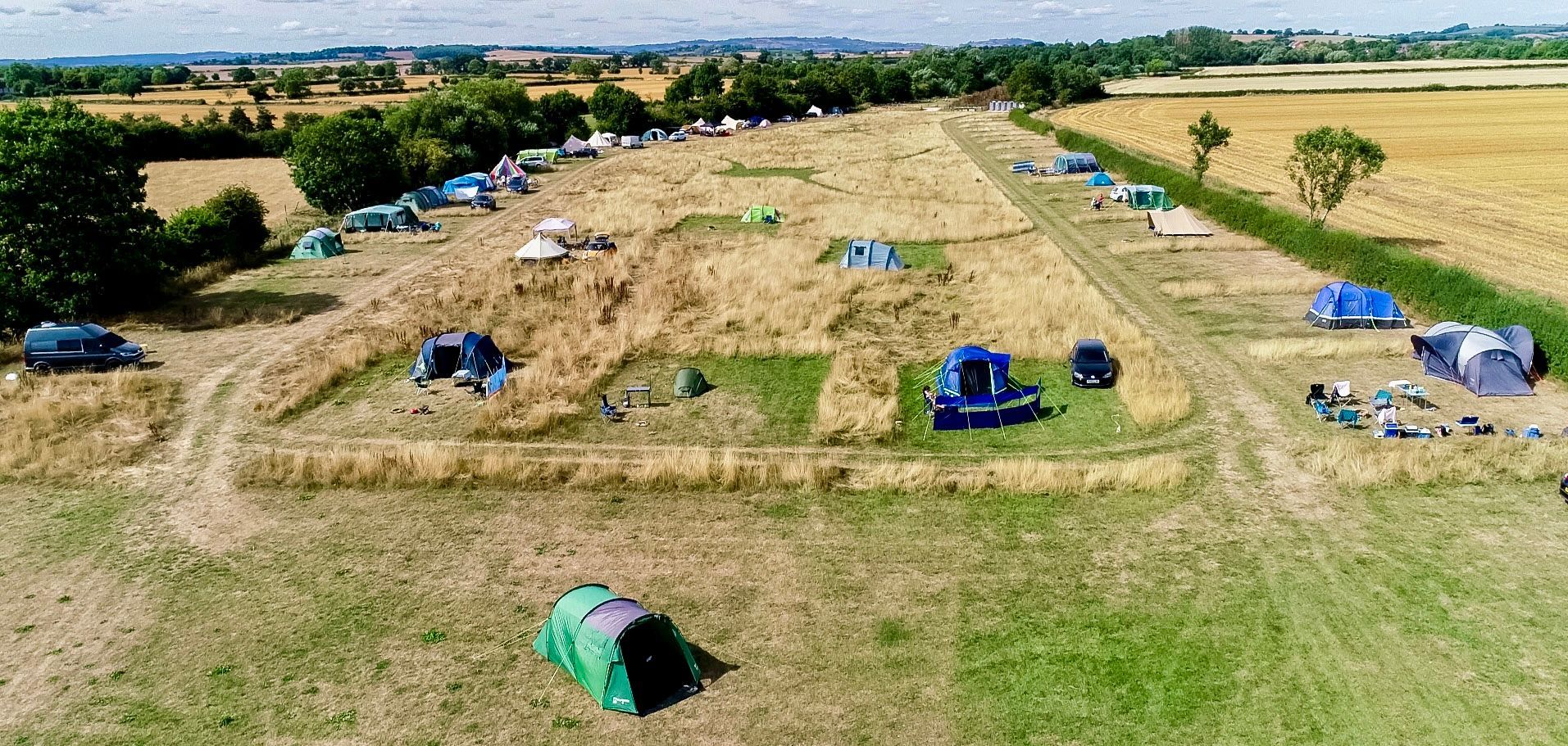 Aerial view of a grassy field with scattered blue tents and shelters near a tree line