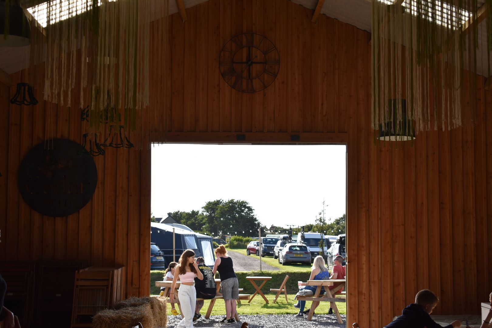 People sitting at tables inside a wooden hall, looking out through a large doorway to a sunny outdoor scene.