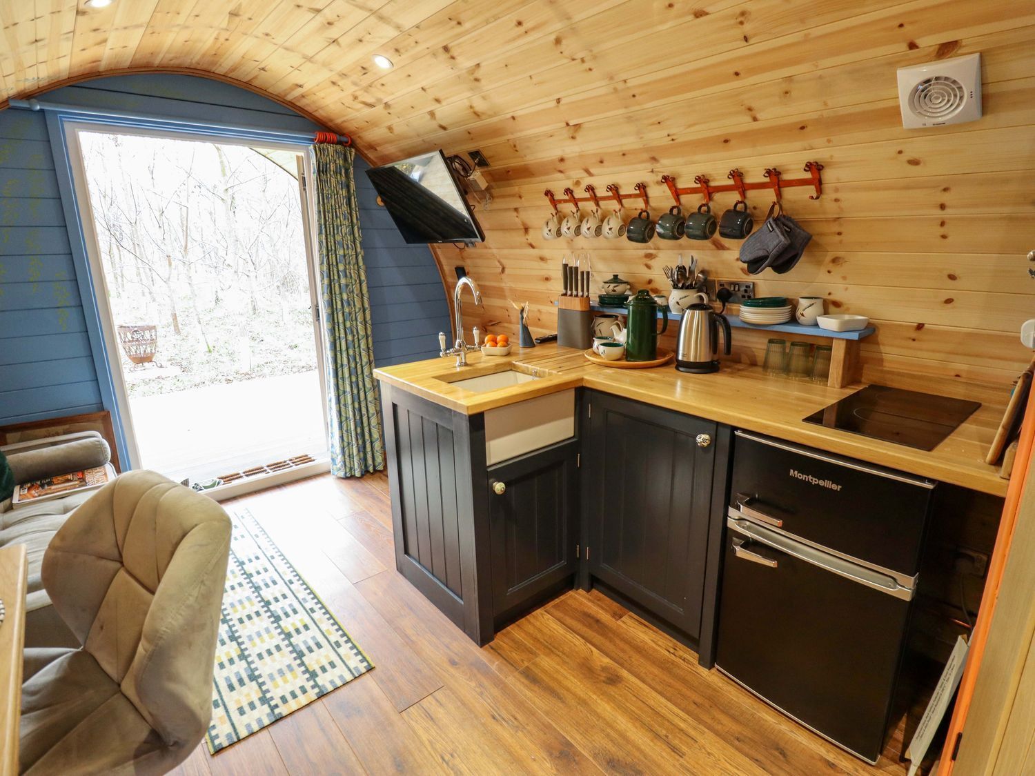 Compact wooden kitchenette with black cabinets, sink, kettle, and open doorway to outside.