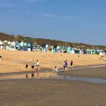 Beach with shallow water, a few people walking, and colorful huts along the shore under a clear blue sky