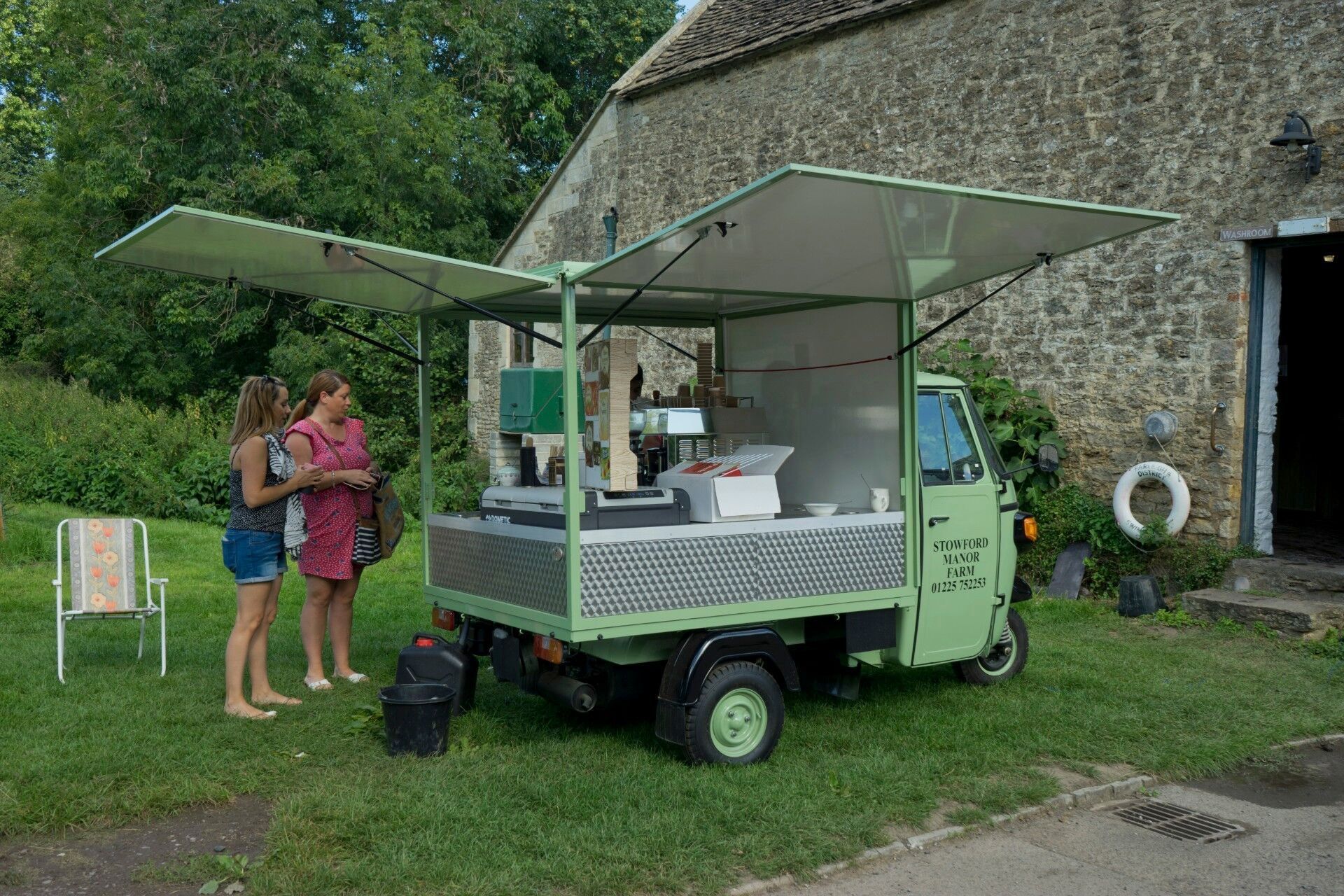 Two people standing by a green food truck with open awning beside a stone building